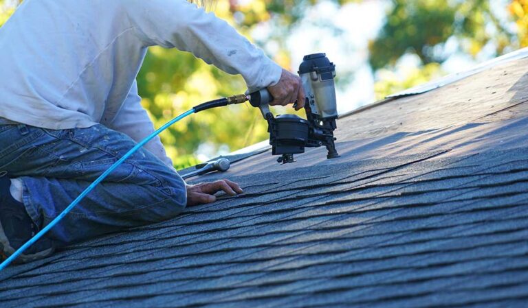 Roofing workers inspect exposed roof decking beside stacked shingles on a residential home
